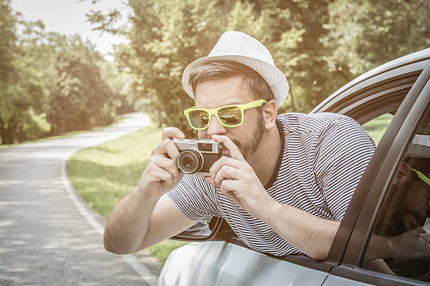 Portrait of young excited tourist guy taking photos through the window from driver’s seat on road trip travel vacation by using vintage retro camera.