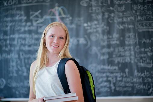 A college student is standing at the front of the classroom in front of the blackboard. She is wearing a backpack and is carrying a stack of books. She is smiling while looking at the camera.