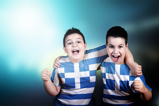 Two adorable boys soccer fans with flag of Greece on t-shirt, embracing celebrate the victory of his team. Happy and shout. Stadium lights in the background. Blue background. Landscape orientation.