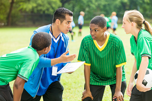 Hispanic soccer coach talks with his coed teen soccer team during time out. He is going over a play with them. The African American male players and the Caucasian female player listen intently to him. The coach gestures as he speaks. The palyers are wearing green jerseys. The female player is holding a soccer ball. The coach is wearing a blue jersey.