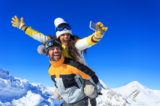 Cheerful young couple hikers making winter selfie on top of the mountain. Both wears warm clothes and gloves, knit hats and sunglasses. Woman in piggyback ride holding the smart phone. Mountain peak and clear sky on background.