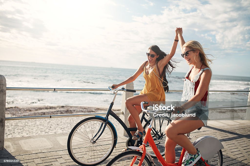 Female friends enjoying cycling on a summer day Female friends enjoying cycling on a summer day. Two young female friends riding their bicycles on the seaside promenade. Friendship Stock Photo