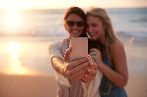 Young female friends on vacation taking selfie on the beach with a smart phone, focus on the hand and mobile phone.