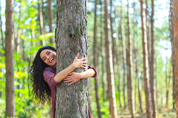Cheerful young woman embracing a tree in the forest Playful young woman embracing a tree in the forest. Standing and looking at camera. Trees and sunbeam on background. hugging tree stock pictures, royalty-free photos & images