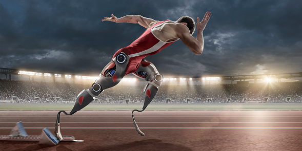 A mid action image of a professional physically disabled male athlete in mid motion, sprinting from starting blocks on an outdoor athletics track. The athlete has two artificial prosthetic futuristic robotic legs and is running in a generic floodlit stadium full of spectators under a stormy evening sky. 