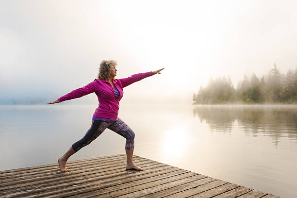 Senior aged woman practicing yoga Senior woman doing morning yoga at a mountain lake warrior position stock pictures, royalty-free photos & images