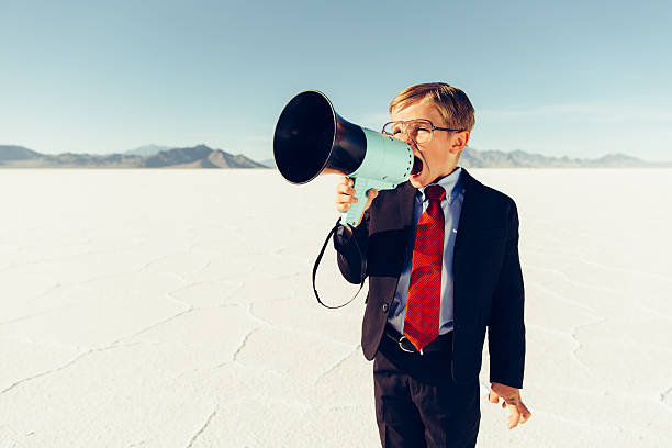 Young Boy Businessman Shouts Through Megaphone A young businessman shouts through a megaphone on the Bonneville Salt Flats in Utah, USA searching for new business opportunities. He is dressed in a suit and red tie with glasses. Retro styling. bonneville salt flats stock pictures, royalty-free photos & images