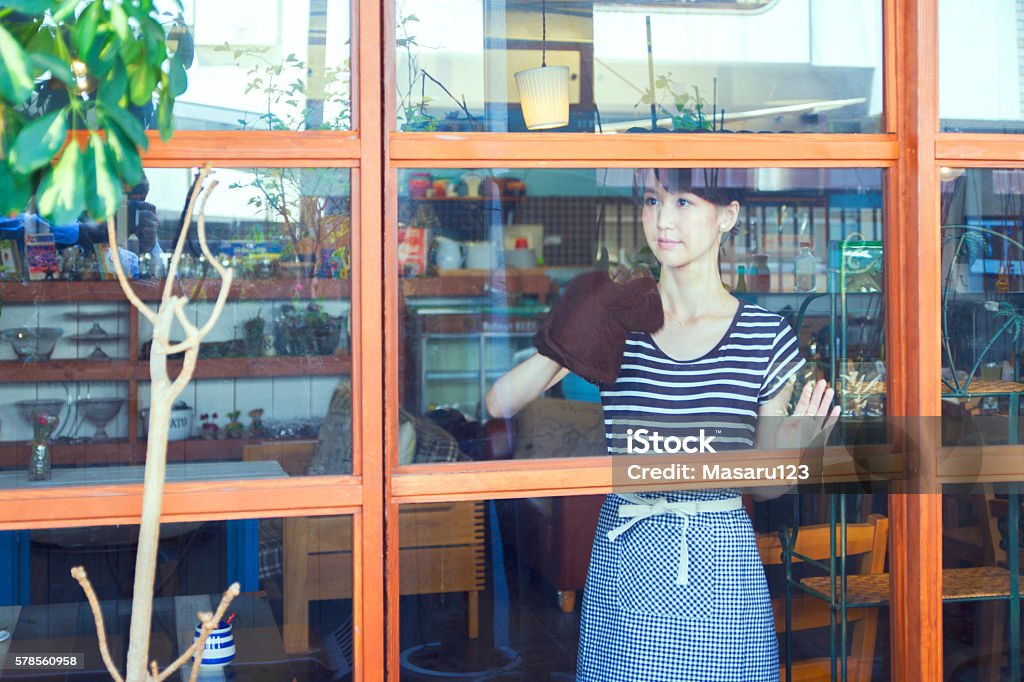 Young woman cleaning a window glass A young woman is cleaning of a window glass at a restaurant (cafe). She wipes a duster on a window glass. She will preparing the shop before business hours. Cleaning Stock Photo