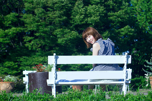 A young woman is sitting on a white bench at a park. She is looking back and smiling.