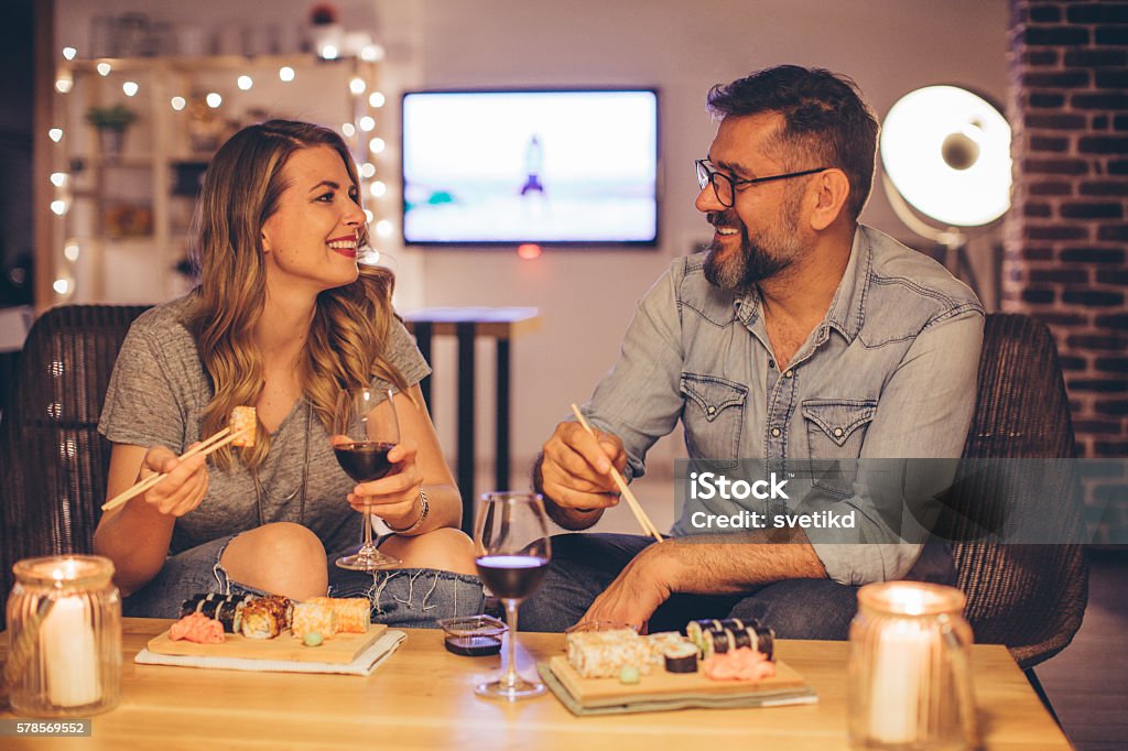 Spending quality time together Two mature people laughing as they sit at a table, drinking vine and eating sushi. Evening or night with beautiful yellow lights lightning the scenes. Love is in the air. Sushi Stock Photo