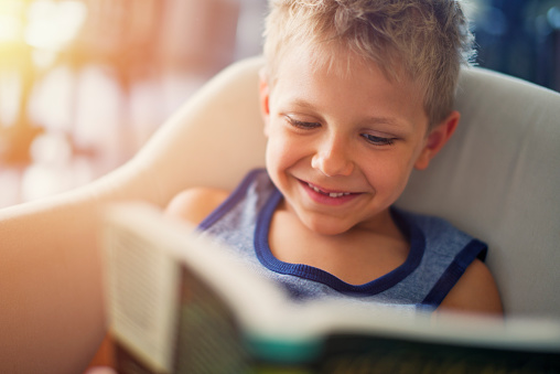 Closeup portrait of a little boy aged 6 reading a book. The boy is sitting in an armchair and is smiling, showing a missing tooth.