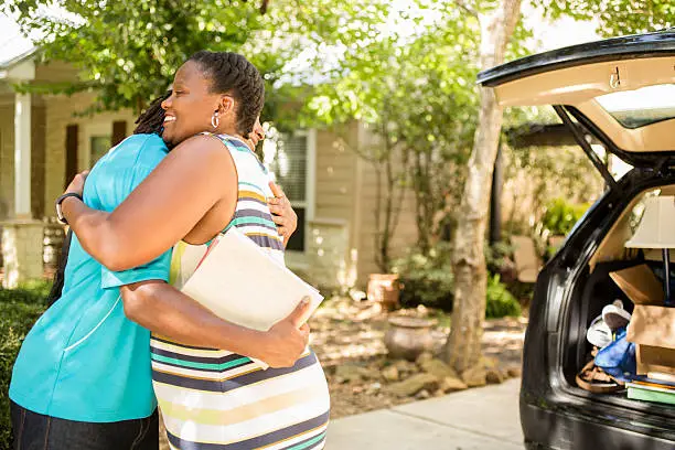 African descent boy heads off to college or moves away from home.  The 18-year-olds' mother is helping him pack up his car as he gets ready for the big move.  He is excited to start his college adventures and gives mom a big hug. He wears a backpack and carries textbooks.  Family events.  Back to school.