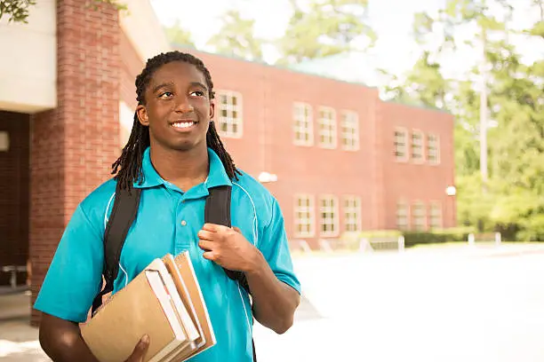 Back to school.   Handsome, African descent boy heads off to college.  The 18-year-old is excited to start his first day of school.  He carries a backpack and textbooks.  College building in background.