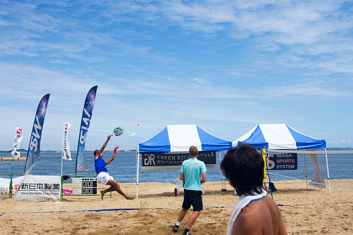 Hakata, Japan - August 22, 2015: A man wearing blue shirt is jumping and hitting a tennis ball in the tennis court. A man playing with him and a man outside of the court are watching his play. International beach tennis tournament in Fukuoka (2015/08/22) 