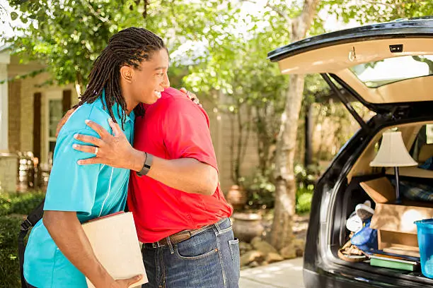 African descent boy heads off to college or moves away from home.  The 18-year-olds' father is helping him pack up his car as he gets ready for the big move.  He is excited to start his college adventures. He wears a backpack and carries textbooks.  Family events.  Back to school.  Hug.