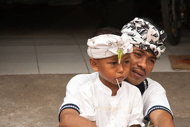 Ubud, Bali, Indonesia - December 8, 2009: A father and his young son both wearing the traditional Balinese ceremonial udeng (headgear). The father is holding the child, who has a lollipop in his mouth.