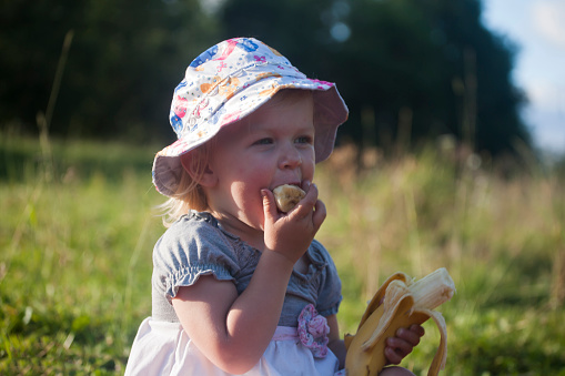 Little girl is eating banana during her walk in sunny day.