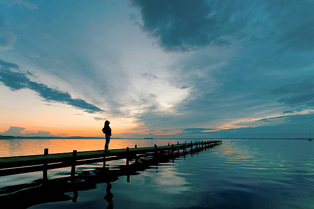 silueta de mujer en lakeside jetty con majestuoso sunset cloudscape - natural looking fotografías e imágenes de stock