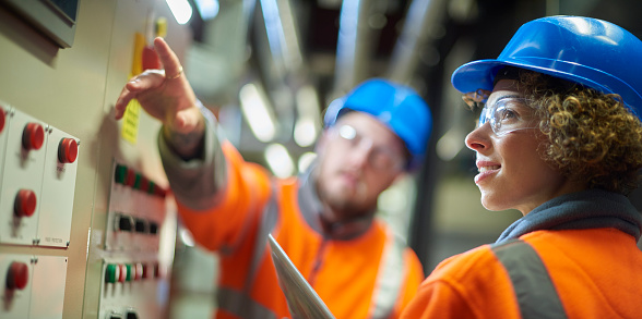 two industrial service engineers conduct a safety check of a control panel and boiler room at a power station . They are both wearing safety equipment and are looking at a control panel .