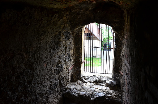 View to outside through small glassless window with grate in the thick stone wall of and old church.