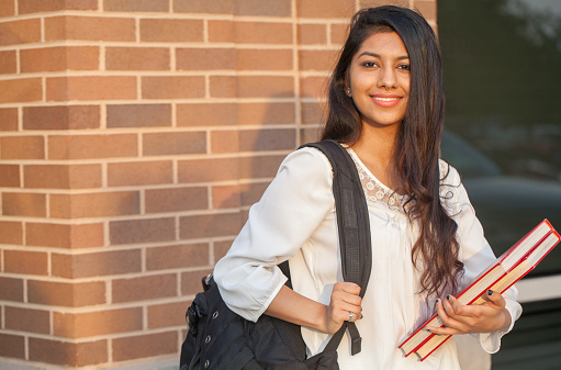 Smiling female young college student of Indian ethnicity carrying backpack and holding books