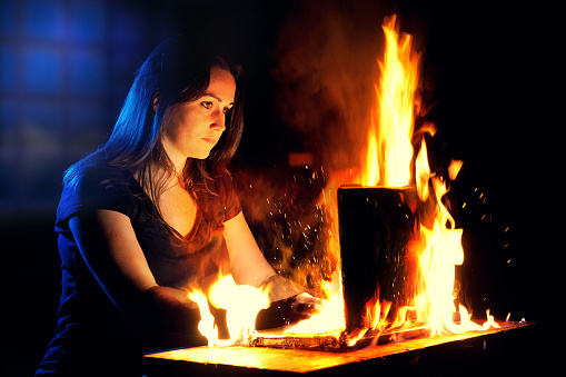 A woman uses a laptop while it is engulfed in flames.