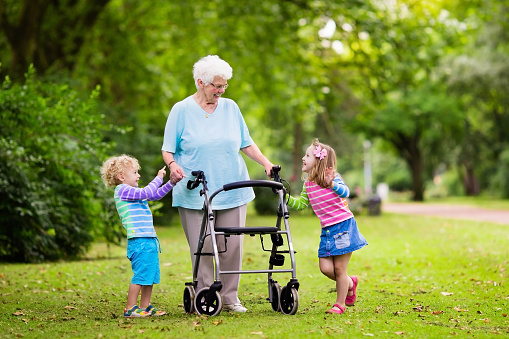 Happy senior lady with a walker holding hands of little boy and girl. Grandmother with grand children enjoy a walk in summer park. Kids supporting disabled grandparent.