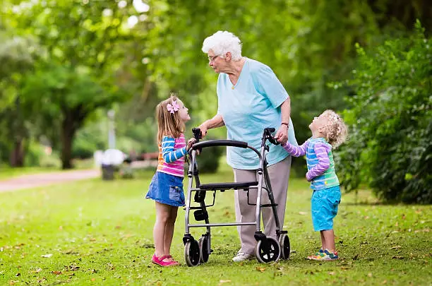 Happy senior lady with a walker holding hands of little boy and girl. Grandmother with grand children enjoy a walk in summer park. Kids supporting disabled grandparent.