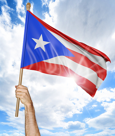 Person holding the national flag of Puerto Rico high in the air against a bright sky and sun rays.