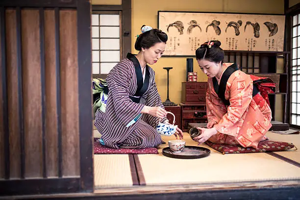 Two Japanese women wearing a kimono drinking tea indoors, in tea room. They are talking and having fun.