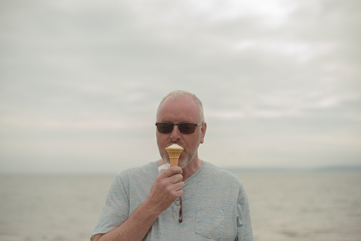 Handsome senior man with sunglasses eating an ice cream cone by the Atlantic Ocean, Hook Head, Wexford, Ireland. 