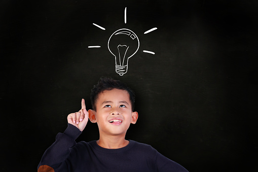Portrait of little Asian student boy smiling and raise his pointing finger up, get an idea gesture, over blackboard with light bulb drawn above his head