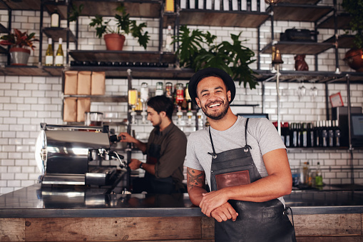 Coffee shop worker smiling to camera, standing at the counter. Happy young man in apron and hat leaning to cafe counter, with waiter working in background.