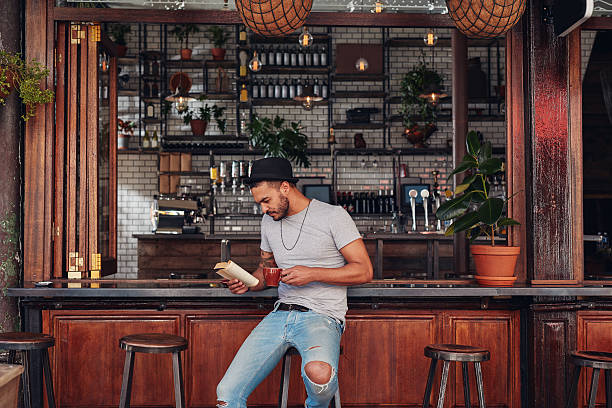 Modern young man reading a book in cafe Portrait of trendy young man sitting at a cafe counter reading a book and drinking coffee cafe culture photos stock pictures, royalty-free photos & images
