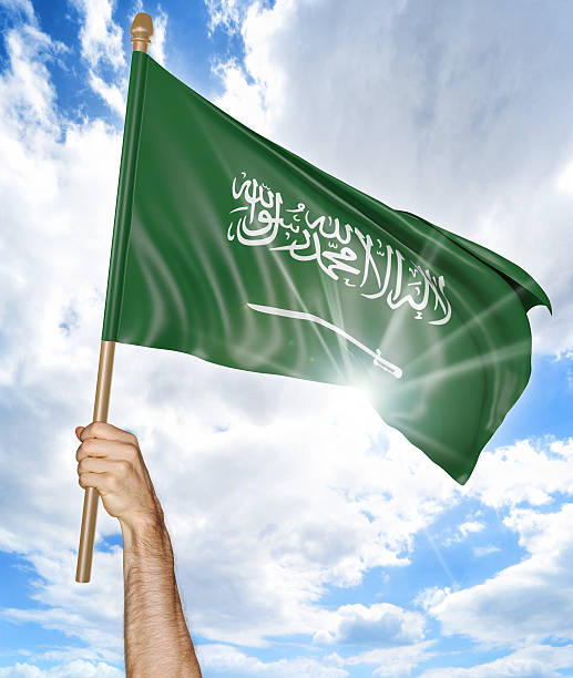 Person holding the national flag of Saudi Arabia high in the air against a bright sky and sun rays.