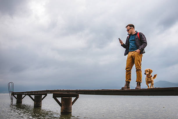 man standing on dock and using smart phone - dog tranquil scene pets animals and pets imagens e fotografias de stock