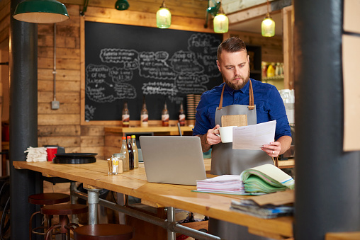 a coffee shop owner checks the delivery notes in his bookkeeping folder on the counter of his busy coffee shop and cross checks them with his online accountancy via his laptop at the end of a busy day .