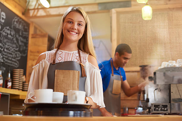 happy young female barista a coffee shop worker smiles to camera as she prepares to take an order to a table in a trendy little coffee outlet . In the background a male colleague is cleaning the coffee machine . first job photos stock pictures, royalty-free photos & images