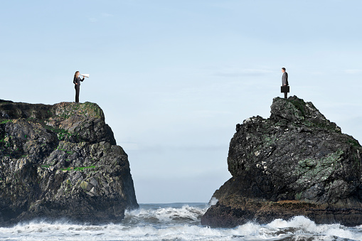 A businesswoman tries to communicate through a megaphone with a businessman as they stand on separate rocky bluffs high above the ocean waves.