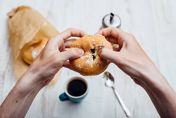 man hands eating bismarck donut with coffee on wooden table - bismarck donuts imagens e fotografias de stock