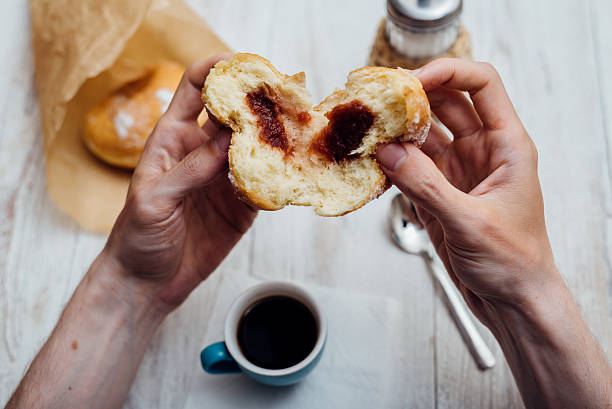 man hands eating bismarck donut with coffee on wooden table - bismarck donuts imagens e fotografias de stock