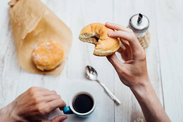 man hands eating bismarck donut with coffee on wooden table - bismarck donuts imagens e fotografias de stock