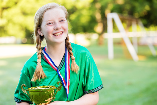 Happy caucasian girl with strawberry blonde hair and freckles. She is laughing and smiling at the camera. She is wearing a green soccer uniform, medal, and braids in her hair. She is standing on the soccer field holding the team trophy.