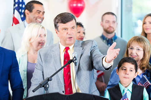 American mature politician in suit standing at tribune and giving a speech during press conference