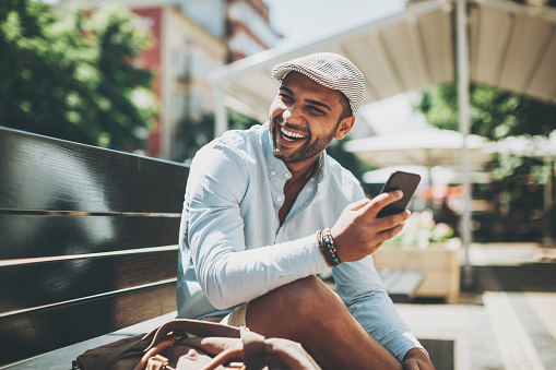 Smiling young East Asian ethnicity man with smart phone sitting on a bench in the city.