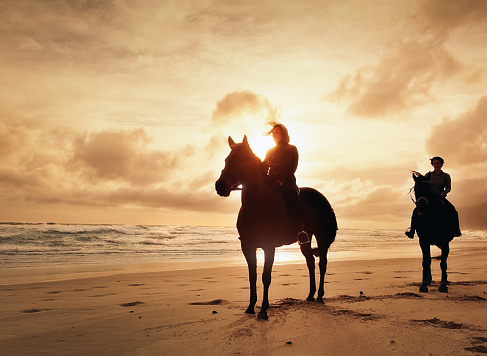 Two women, riding horses on a  beach on a windy winter's afternoon, are almost silhouetted against the choppy sea and cloudy, gold-tinged sky as the sun begins to set.