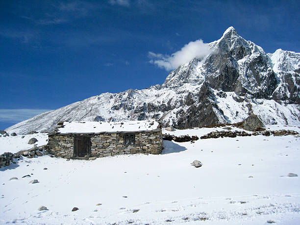 Everest Region, Nepal - March 17, 2010: A  stone cottage with snow-covered roof and snow mountain behind has been seen during the everest base camp trekking route in bright blue sky.