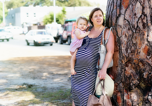 Overtired and overburdened young mother, very pregnant with her second child, leans against a tree, carrying her toddler daughter and many shopping bags.