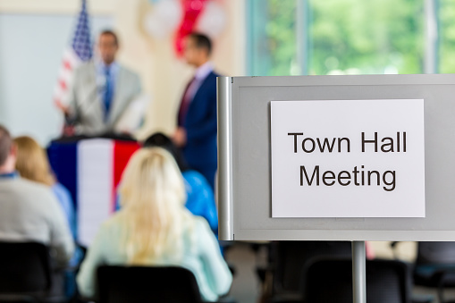 Town hall meeting sign with the town hall meeting proceeding in the background. There is an american flag that can be identified.