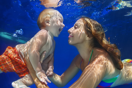 Funny happy family - mother, baby son learn to swim, dive underwater with fun in pool. Healthy lifestyle, active parent, people water sports activity and swimming lessons on summer holiday with child